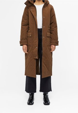 Brązowy pikowany długi płaszcz z zatrzaskami i suwakiem, noszony otwarty na białą koszulę, czarne szerokie spodnie o długości do kostek oraz czarne botki.