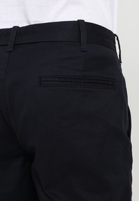 Námorně modré kalhoty s plochou zadní kapsou, vyrobené z texturované látky. Obsahují poutko na opasek v oblasti pasu a mají hladký povrch.