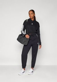 Giovane donna in tuta nera Adidas, sneakers bianche, che porta una borsa da palestra nera sulla spalla, in piedi di fronte a uno sfondo semplice.