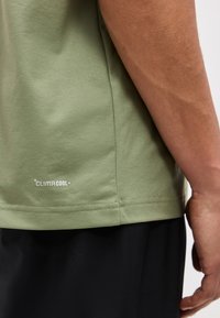 Πράσινο πουκάμισο με το λογότυπο "Climacool" κοντά στο τελείωμα, φορεμένο από ένα άτομο που στέκεται με το δεξί του χέρι χαλαρό δίπλα σε μαύρο παντελόνι.