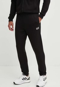 Pantaloni sportivi neri con una texture liscia, dotati di un logo EA7 sulla coscia sinistra, vita elastica e gambe affusolate. Indossati con sneaker bianche.