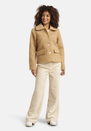 Tan wollen jack met een gestructureerde afwerking, grote kraag, vo pockets en knoopsluiting, gecombineerd met crèmekleurige broek met wijduitlopende pijpen en sneakers.