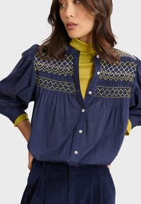 Camicia blu navy con bottoni, adornata da un intricato ricamo giallo e bianco sul petto, caratterizzata da maniche a sbuffo e una vestibilità morbida.