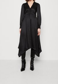 Robe noire longue avec un décolleté en V, un corsage plissé et un ourlet asymétrique fluide. Fabriquée dans un tissu lisse, elle possède des manches longues et des poignets structurés.