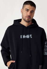 Černá mikina s kapucí, která má texturovaný potisk na přední straně v modré barvě, boční kapsy a žebrované manžety. Obsahuje branding na jedné rukávu.