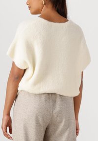 Witte, fuzzy trui met korte mouwen en een ronde halslijn, gecombineerd met beige broeken in een visgraatpatroon. Zachte textuur, ontspannen pasvorm.