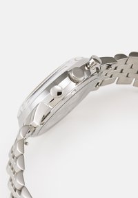 Montre en métal argenté avec une finition polie, boutons chronographe et bracelet en métal avec des maillons entrelacés.