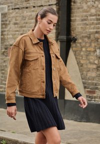 Brun ruskindsjakke med knaplukning foran, to brystlommer og en afslappet pasform, kombineret med en navyblå ribstrikket kjole.