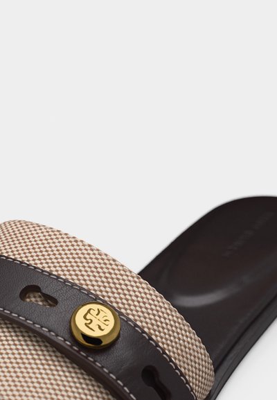 Κοντινό πλάνο μιας μπεζ υφασμάτινης σαγιονάρας με λουρί από σκούρο καφέ δέρμα που φέρει ένα χρυσό κυκλικό έμβλημα.