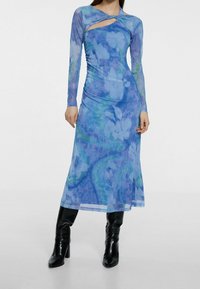 Blaues Kleid mit floralem Muster, langen Ärmeln, mit Ausschnitt-Design, schlanker Passform und fließendem Silhouette, kombiniert mit knielangen schwarzen Stiefeln.