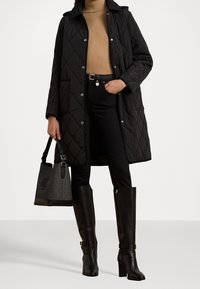 Svart vadderad lång kappa med knäppknappar, bärs över en beige tröja och svarta jeans. Svarta knähöga stövlar och en grå handväska kompletterar looken.