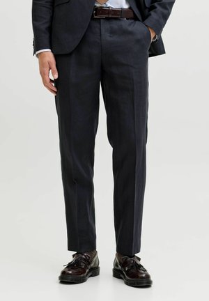 Homme portant un pantalon sombre sur mesure, une ceinture en cuir marron et des mocassins à pampilles marron foncé, debout avec une main dans la poche.