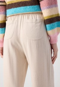 Pantaloni bej, texturat, cu un tiv elastic și două buzunare la spate. Țesătura prezintă un model subtil și un finisaj moale.