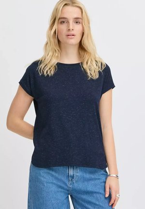 Femme blonde portant un t-shirt bleu marine à manches courtes et un jean bleu clair, debout avec une main derrière le dos.