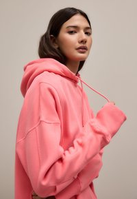 Prevelik pulover s kapuco v svetlo roza barvi. Ima vrvico za zategovanje in rebraste manšete. Mehka tkanina z gladko teksturo.