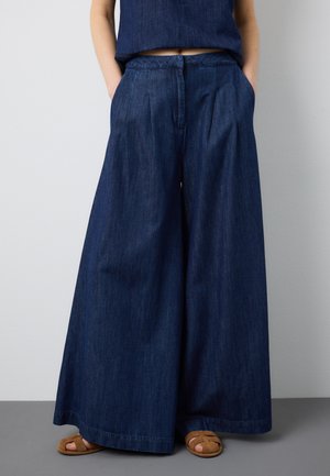 Person trägt dunkelblaue Jeans mit weitem Bein, die Hände in den Taschen und braune Sandalen mit offenem Zehenbereich, steht vor einer schlichten Wand.