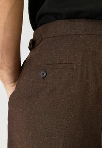 Pantalons marron en tissu texturé avec un motif subtil, dotés d'une poche arrière et d'un détail de bouton noir à la taille.