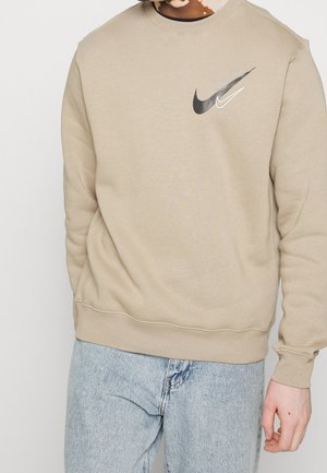 Beiges Sweatshirt mit schwarzen und weißen Nike-Logos und dem Text "Just do it", getragen von einer Person mit Vitiligo-Haut zusammen mit hellblauen Jeans.