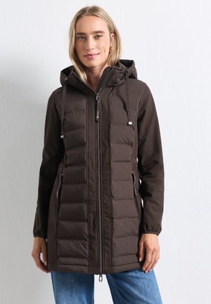 Frau trägt eine dunkelbraune gesteppte Kapuzenjacke mit Reißverschlusstaschen und blaue Jeans, steht vor einer schlichten hellen Wand.