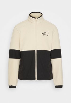 Veste en polaire beige et noire avec une fermeture éclair à l'avant ; portée avec un short en velours côtelé marron et des baskets blanches avec des accents rouges et bleus.