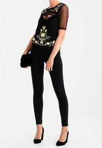 Blusa nera trasparente con ricamo floreale, abbinata a leggings neri. Modella che tiene una piccola clutch nera. Tacchi neri completano l'outfit.