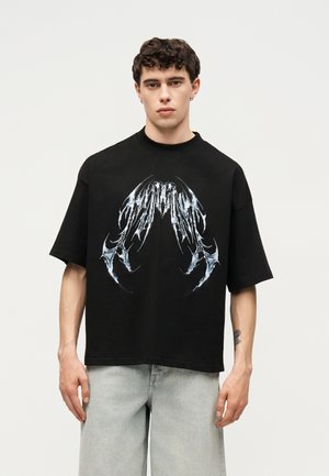 Jeune homme portant un t-shirt noir ample avec un grand motif abstrait de griffe argentée, un jean gris clair et des boucles d'oreilles créoles argentées.