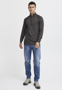 Mørkegrå zip-neck genser med teksturert stoff, ribbestrikkede mansjetter og kant, kombinert med lyseblå jeans og grå sneakers.