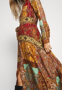 Vrouw in een vloeiende lange mouw jurk met een intricaat paisley- en patchworkpatroon in rood, oranje, geel en turquoise.