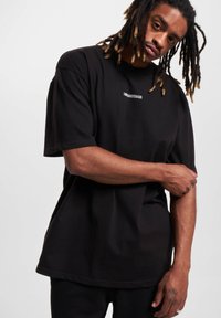 Juodas per didelis T-palaidinukas, pagamintas iš medvilnės, su trumpomis rankovėmis ir giliu apykaklės kraštu. Subtilus balto logo detalė krūtinėje.