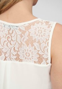 Haut blanc avec un dos en dentelle décoré de motifs floraux, texture transparente et tissu avant lisse. Encolure ronde et sans manches.