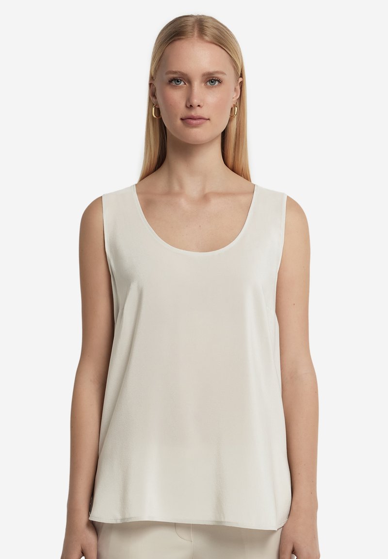 Femme blonde portant un top crème sans manches avec un décolleté rond et des boucles d’oreilles créoles dorées sur fond blanc.