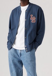 Chaqueta sudadera azul marino con cuello redondo, botones de presión, logo bordado "LS" y bolsillos laterales, usada sobre una camisa blanca y jeans claros.