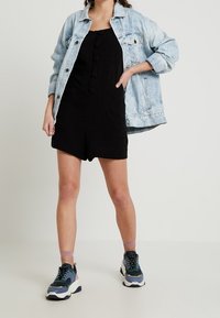 Black romper with front buttons, layered under a light blue denim jacket. Paired with chunky sneakers featuring teal, gray, and white accents.