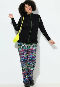 Sort sweater med grøn ruflede kant, parret med farverige mønstrede bukser med tekst i pink, blå og gul. Sorte sko og gul taske.