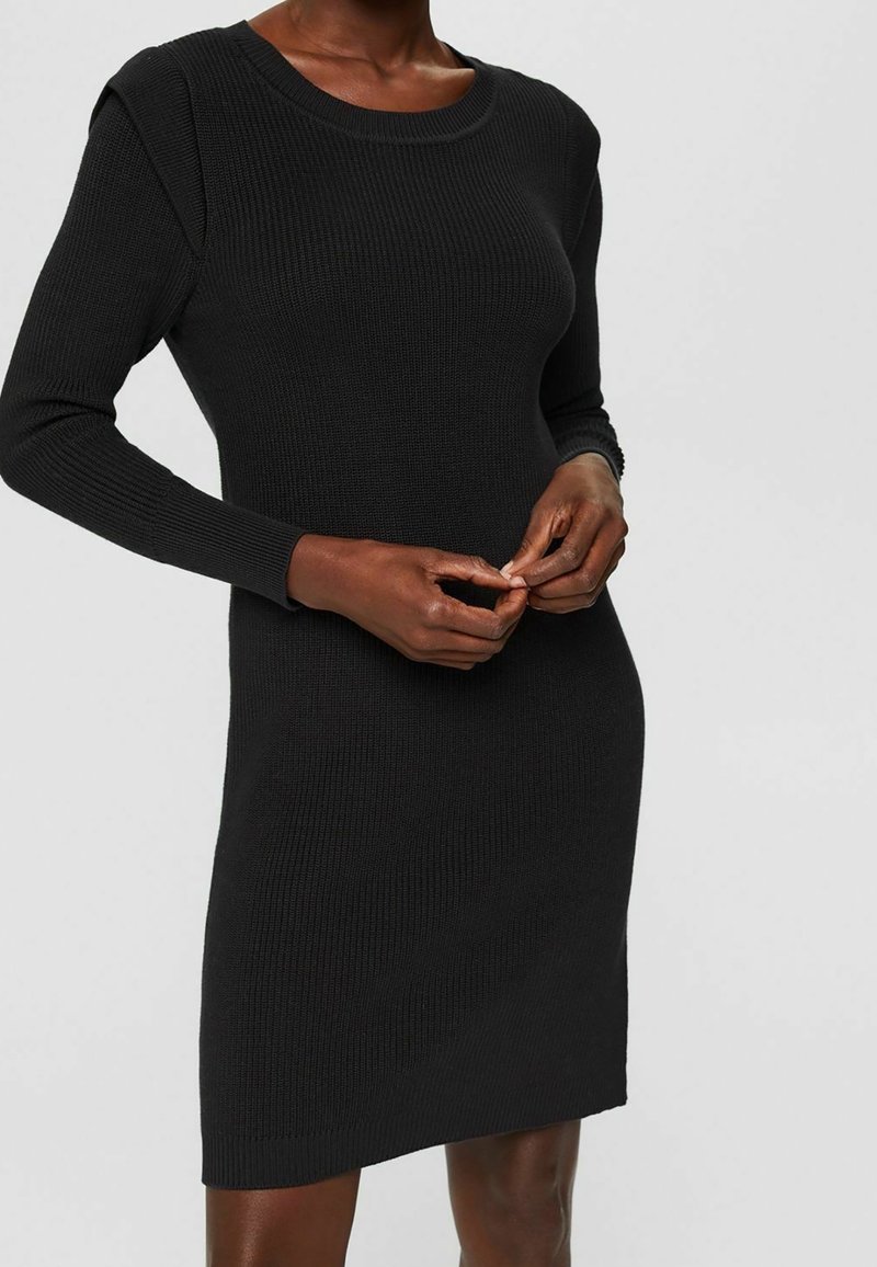Femme portant une robe noire moulante en maille côtelée à manches longues, mains délicatement jointes à la taille, sur un fond gris uni.