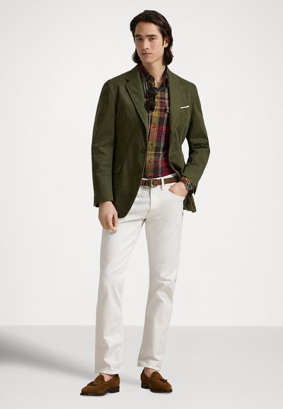 Πράσινο σακάκι πάνω από καρό πουκάμισο, λευκό παντελόνι και καφέ σουέτ παπούτσια. Φοράει γυαλιά ηλίου και ρολόι, με καθαρές γραμμές και εφαρμογή ραμμένη στα μέτρα.