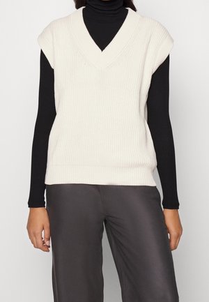 Personne portant un gilet sans manches blanc en tricot par-dessus un col roulé noir à manches longues et un pantalon gris foncé, debout les bras détendus.