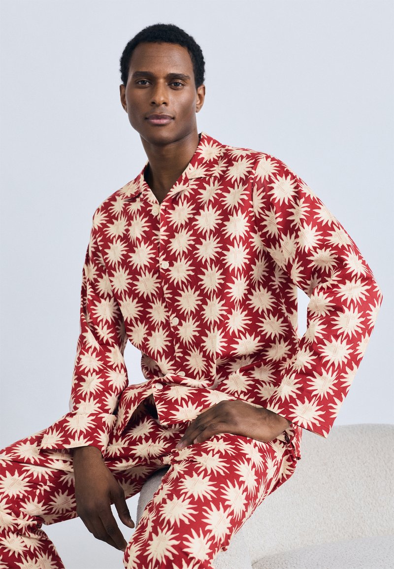 Set de pijama roșu cu un model de stelă în crem. Bluza are guler și mâneci lungi, iar pantaloni asortați completează ținuta.