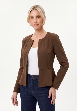Femme blonde portant des boucles d’oreilles en or, un blazer marron ajusté, un haut blanc et un jean bleu foncé, debout devant un fond clair uni.