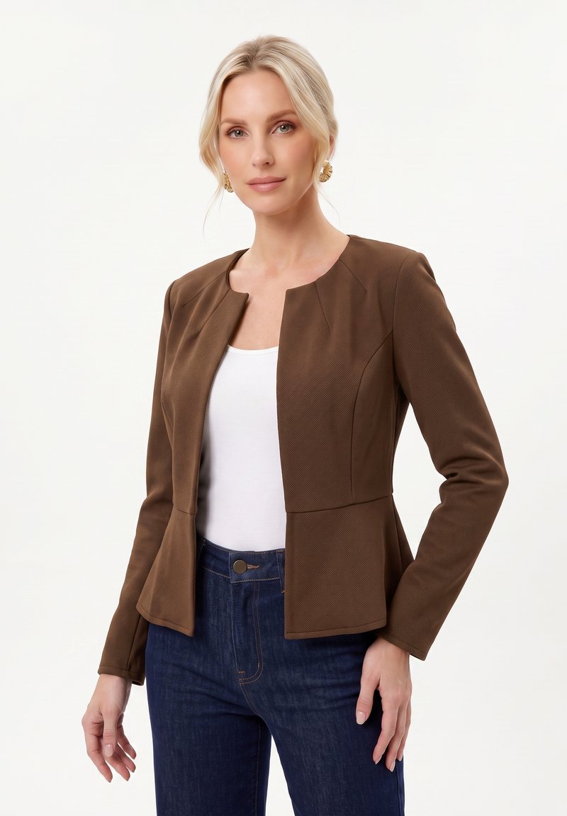 Femme blonde portant des boucles d’oreilles en or, un blazer marron ajusté, un haut blanc et un jean bleu foncé, debout devant un fond clair uni.