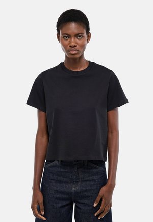 Femme aux cheveux courts portant un t-shirt noir uni à manches courtes et un jean bleu foncé, debout devant un fond gris clair.