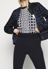 Schwarze Daunenjacke, gemustertes Sweatshirt in Schwarz und Weiß mit "TOMMY"-Text und schwarze Leggings. Glatte Texturen, figurbetontes Design.