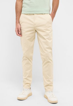 Beige Baumwollhosen mit schmaler Passform, ausgestattet mit Seitentaschen, flachem Bund und leichter Textur, kombiniert mit weißen Sneakern.