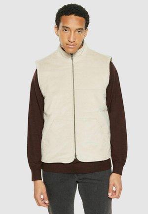 Mann trägt beige zip-up ärmellose Weste über einem dunkelbraunen langärmligen Hemd und dunkler Hose, steht vor einem einfarbigen hellen Hintergrund.