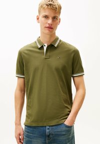 Oliwkowa koszulka polo z białym i granatowym paskowanym kołnierzykiem. Krótkie rękawy, strukturalna tkanina i małe logo na lewej piersi. Casualowy krój.