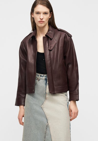 Γυναίκα με σκούρο καφέ δερμάτινο μπουφάν πάνω από μαύρο τοπ και δίχρωμη τζιν φούστα, στέκεται μπροστά σε ένα απλό φωτεινό φόντο.