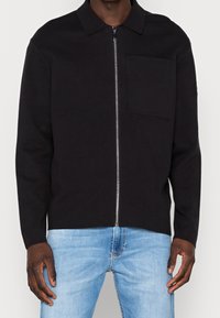 Svart zip-up jacka i stickad kvalitet, med krage, en enda bröstficka och långa ärmar, tillsammans med ljusblå jeans.