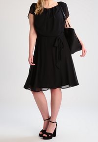 Černé šaty ke kolenům z měkké, splývavé tkaniny. Mají krátké rukávy, pásek v pase a volný střih. V kombinaci s černými sandály na blokovém podpatku.