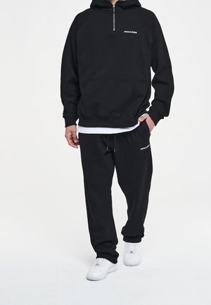 Persona che indossa una felpa nera con mezza zip e pantaloni della tuta neri coordinati, con sneakers bianche, in piedi davanti a uno sfondo bianco semplice.