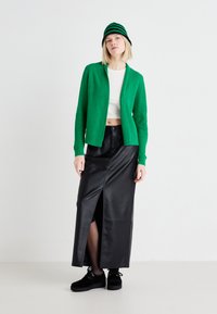 Cardigan côtelé vert sur un crop top blanc, associé à une jupe maxi en cuir noir avec une fente avant et des chaussures à plateforme noires.